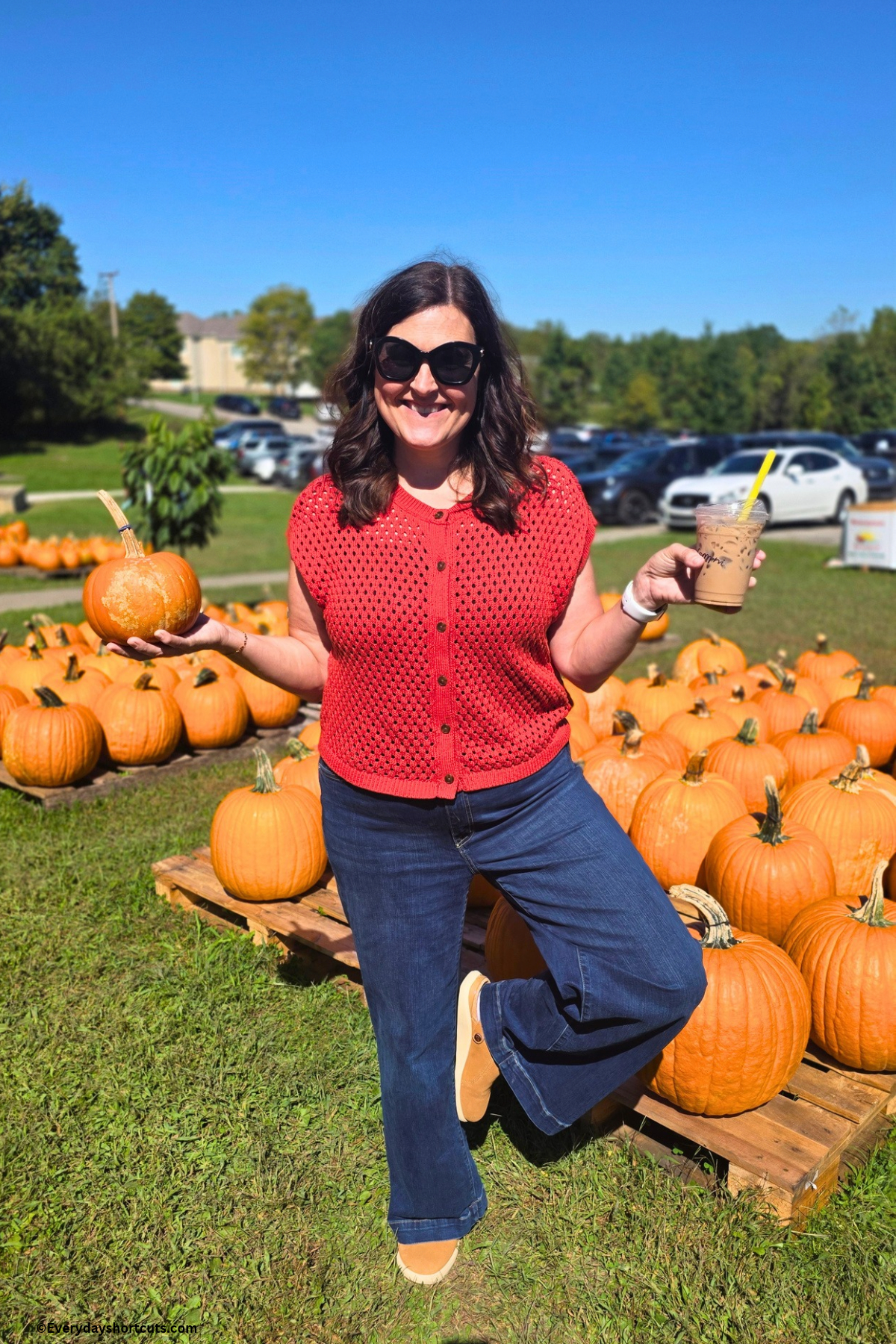 girl at pumpkin patch standing on one leg and holding an iced coffee in one hand and a little pumpkin in the other hand