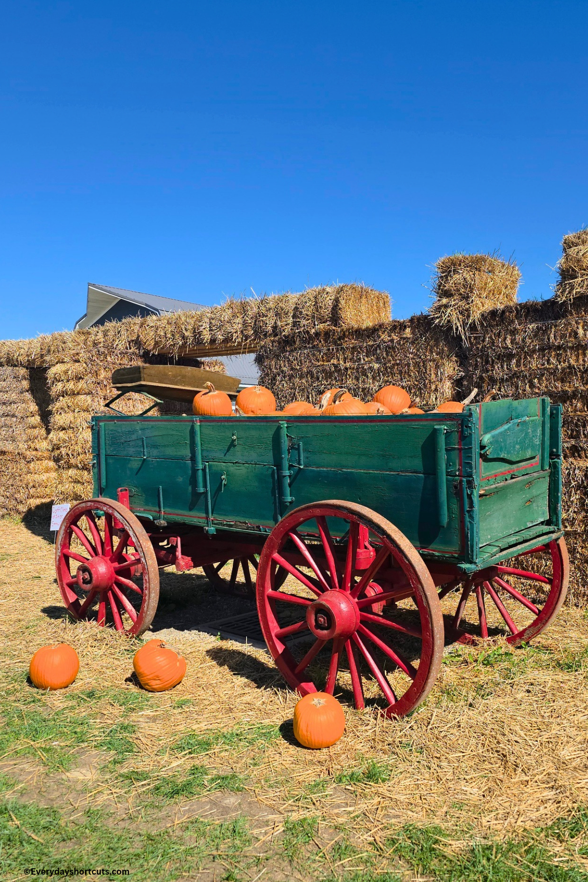 a green wagon with red wheels holding pumpkins next to a straw castle
